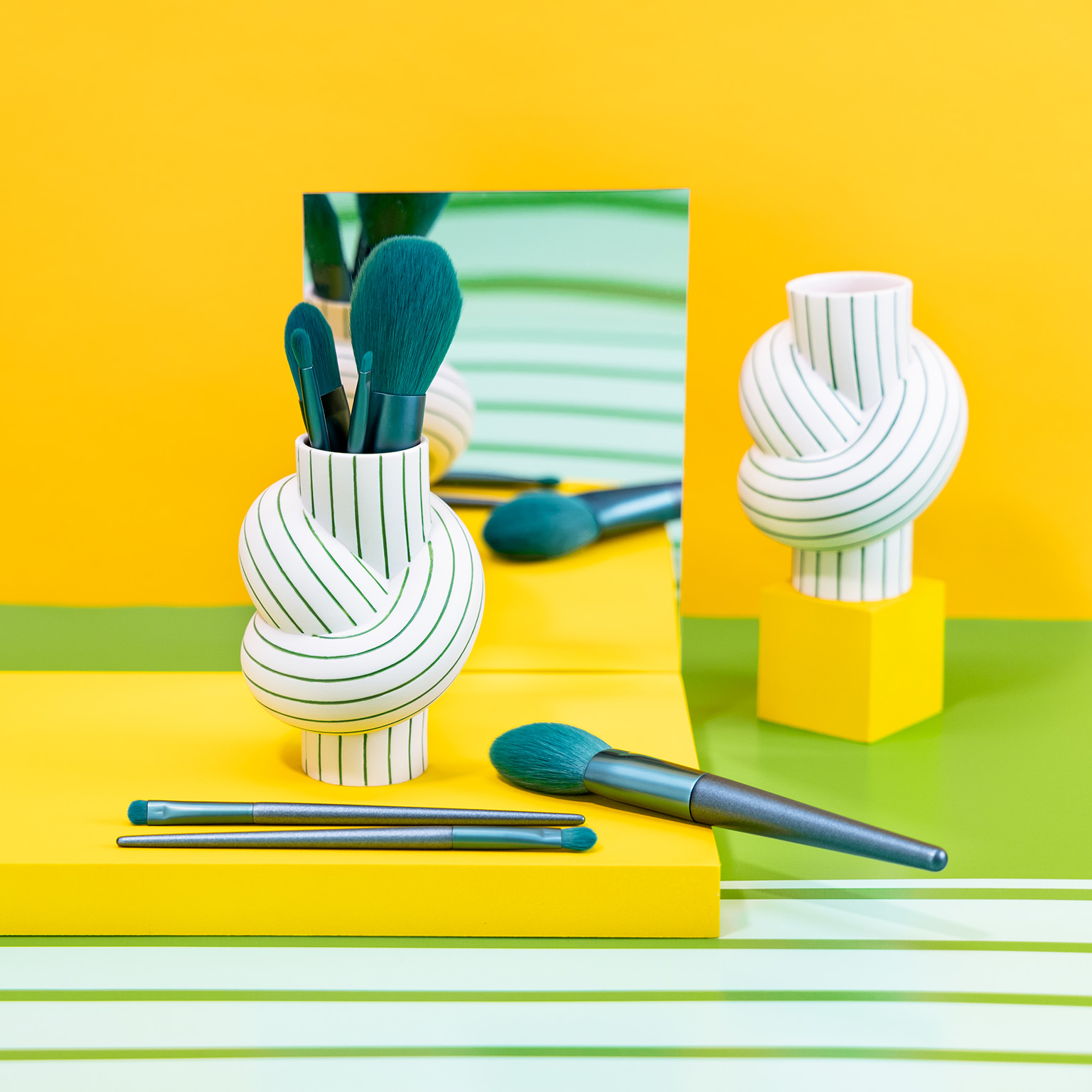 Vase Node Stripes Apple filled with petrol-coloured make-up brushes against a yellow background with white and green stripes and mirror.